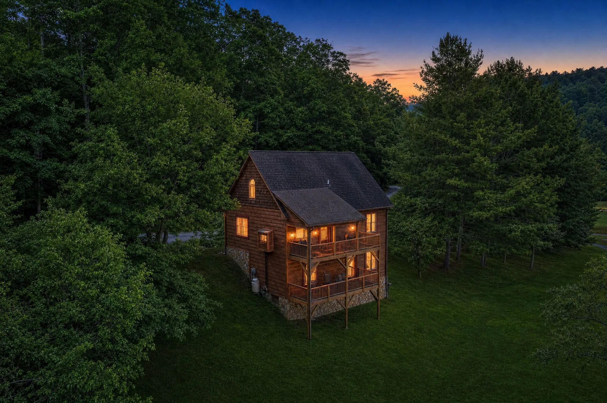 The cedar-shake cabin at dusk with warm-lit windows and a summer sunset behind the trees